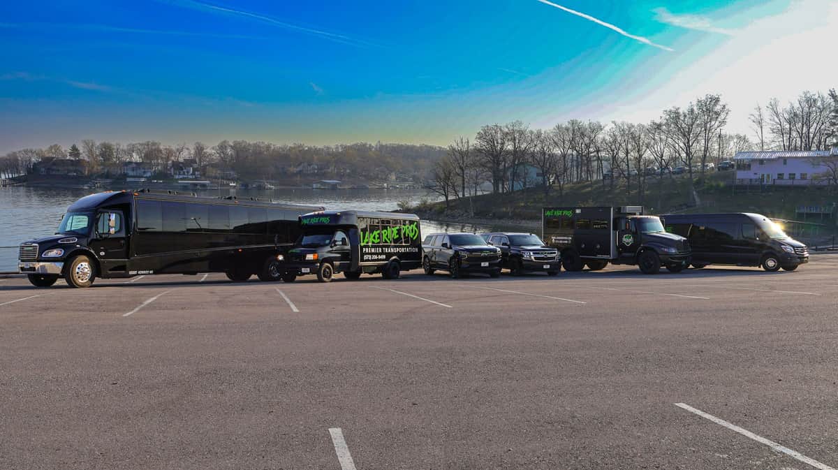 Lake Ride Pros luxury transportation fleet lined up at Lake of the Ozarks including limo bus, Rescue Squad truck, SUVs, and Mercedes Sprinter vans with lake view in background